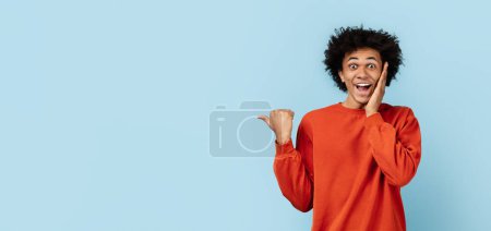 chico afroamericano en suéter naranja mirando sorprendido y apuntando hacia los lados, fondo azul aislado, espacio de copia