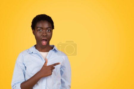A surprised African American woman with an amusing expression, pointing to her side, isolated on a vivid yellow background symbolizing surprise and spontaneity of zoomers