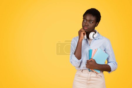 Pensive young black woman student holds her chin, contemplating next steps with her notebooks, a trait of the inquisitive zoomer, against an isolated yellow background, copy space