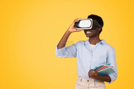 Afroamerikanerin steht vor leuchtend gelbem Hintergrund, das Gesicht von einem Virtual-Reality-Headset verdeckt, während sie bunte Bücher in der Hand hält