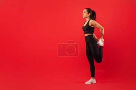 A woman in athletic wear is standing on one leg and holding her other foot behind her, stretching her quadriceps muscle. She maintains a focused expression, demonstrating proper form for a leg stretch
