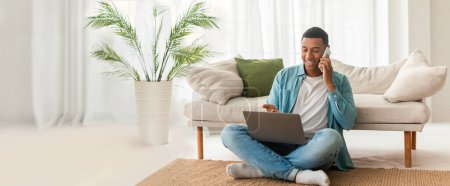 Photo for Relaxed African American guy using laptop and chatting on a phone at home, web-banner format with copy space - Royalty Free Image