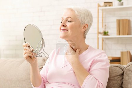 Photo for Senior woman is seated on a couch, holding a mirror in her hand. She appears to be examining her neck intently. The room is dimly lit, with soft furniture in the background. - Royalty Free Image