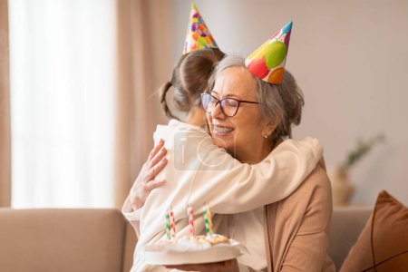A tender moment captured as an older woman embraces a young girl, both wearing celebratory birthday hats. The affection and joy between them are evident in the heartfelt hug shared.