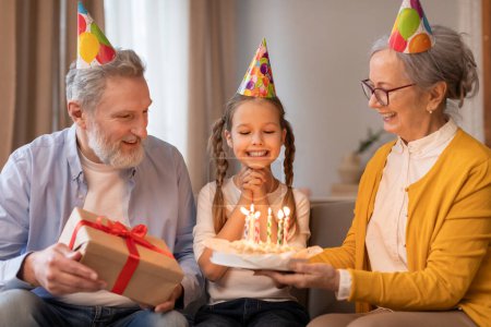 Foto de Una joven con una sonrisa chispeante se sienta entre sus adorables abuelos, todos vistiendo sombreros de fiesta festivos, la abuela sostiene el pastel de cumpleaños con velas encendidas, el abuelo presenta un regalo - Imagen libre de derechos