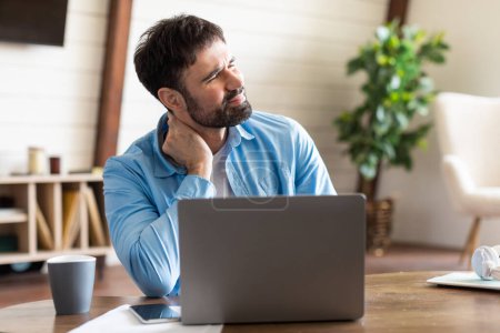 Photo for A man is sitting at a wooden table, focused on his laptop with a pained expression, rubbing the back of his neck. The setting appears to be a bright and cozy home office - Royalty Free Image