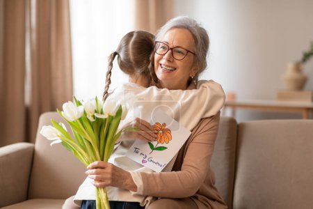 A heartwarming scene unfolds as a young girl with braided hair embraces her smiling grandmother, presenting her with a bouquet of fresh white flowers and a colorful handmade card.