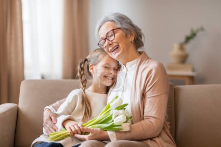 Téléchargez les photos : Une femme âgée joyeuse avec des lunettes étreint chaudement sa petite-fille, qui tient un bouquet de tulipes blanches fraîches, intérieur de la maison - en image libre de droit