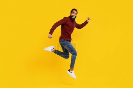 Indian man wearing a red sweater is captured mid-jump in the air. His arms are raised, and his legs are bent as he springs upwards. The background is yellow, emphasizing the mans movement.