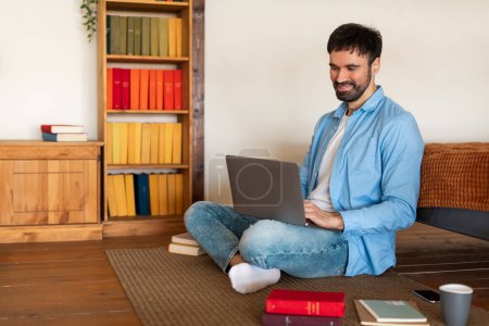 Ein Mann sitzt auf dem Boden und tippt auf einem Laptop vor sich. Er wirkt fokussiert und beschäftigt mit dem Laptop-Bildschirm, umgeben von einer einfachen Inneneinrichtung.