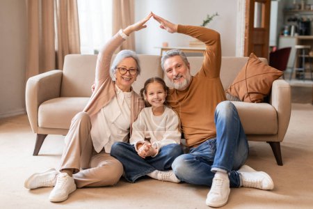A smiling elderly couple and a young girl sit on the floor in a cozy living room, forming a house outline with their hands above them, symbolizing unity and family love.