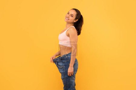 Eine fröhliche junge Frau steht vor gelbem Hintergrund und hält ihre übergroßen Jeans in der Hand, um ihre erfolgreiche Gewichtsabnahme zu zeigen. Ihr strahlendes Lächeln und ihre selbstbewusste Pose vermitteln ein Erfolgserlebnis.