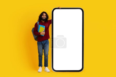 Hombre indio alegre con un estudiante de barba se para al lado izquierdo, sosteniendo libros coloridos y apoyándose en un teléfono inteligente masivo de pantalla en blanco