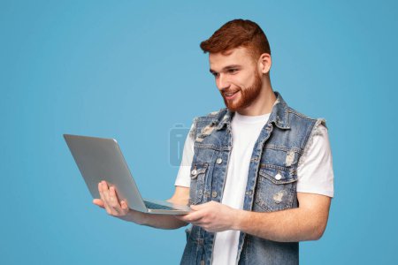 Smiling millennial redhaired man having video conversation on laptop on blue studio background, copy space