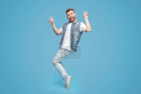 Photo for Awesome guy showing peace gesture and standing on tiptoes on blue background, copy space - Royalty Free Image