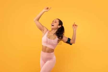 Foto de Una mujer con un atuendo deportivo rosa golpea el aire con emoción, vistiendo un brazalete deportivo negro, usando auriculares inalámbricos, escuchando música, contra un animado telón de fondo amarillo. - Imagen libre de derechos