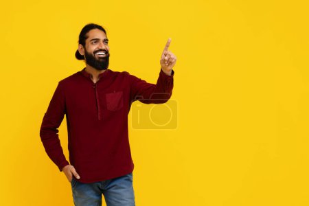 Foto de Hombre alegre indio con barba está de pie sobre un fondo amarillo brillante, apuntando hacia arriba con su mano izquierda, tocando la pantalla virtual invisible - Imagen libre de derechos