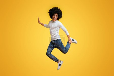 Photo for Joyful bushy black guy having fun, jumping on one leg in air, orange studio background - Royalty Free Image