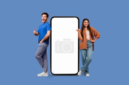 Young man and woman pose beside a large smartphone with white screen mockup copy space on a blue background