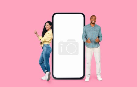 Emocionada pareja Multiracial haciendo un gesto positivo al lado de una pantalla de teléfono inteligente en blanco sobre un fondo rosa