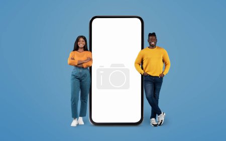 Foto de Hombre y mujer afroamericanos con ropa vibrante posando junto a una pantalla de teléfono inteligente en blanco sobre fondo azul - Imagen libre de derechos