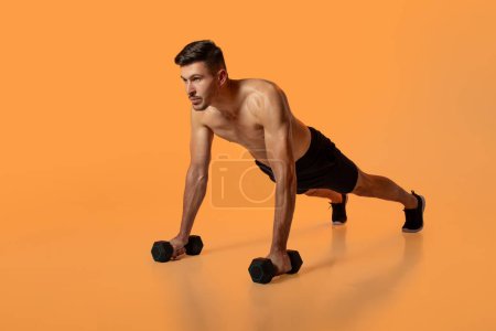 Foto de Un hombre está ejecutando un ejercicio push-up en el suelo de un gimnasio mientras sostiene dos pesas en sus manos. Él está enfocado y manteniendo la forma adecuada mientras baja y levanta su cuerpo de una manera controlada. - Imagen libre de derechos