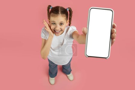 Téléchargez les photos : Une jeune fille aux cheveux tressés tient un smartphone avec un écran vierge et des gestes, sur fond rose, vue de dessus - en image libre de droit