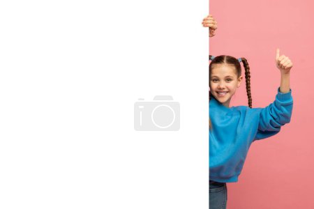 Foto de Una joven alegre con el pelo trenzado en un suéter azul está detrás de una pancarta blanca en blanco, da un signo de pulgares hacia arriba sobre un fondo rosa partido - Imagen libre de derechos