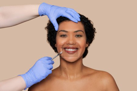 A smiling brazilian woman with curly hair is being carefully attended to by a professional in blue gloves, who is administering a cosmetic injection on her lower lip