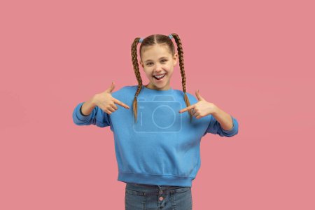 Jovencita alegre con coletas en un suéter azul apuntando con confianza a sí misma sobre un fondo rosa