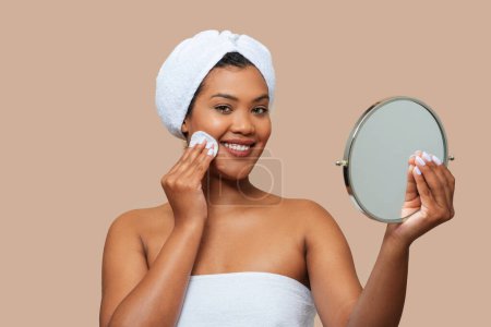 A young, cheerful woman with a towel wrapped around her hair is gently applying facial toner to her cheek. She holds a small, round mirror in one hand to ensure precise application