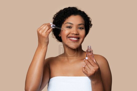 A cheerful brazilian woman with curly hair is holding a small bottle of facial serum and applying the product to her face with a dropper.