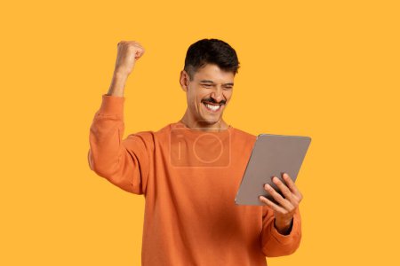 Foto de Un hombre con un suéter naranja es visto sosteniendo una tableta en sus manos. Aparece enfocado en la pantalla, involucrado en actividades de tableta. - Imagen libre de derechos
