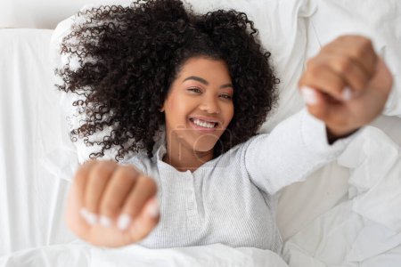 Photo for A cheerful young woman with curly hair is lying on her back in a comfy bed, stretching her arms towards the camera with a relaxed and happy expression, suggesting she just woken up. - Royalty Free Image
