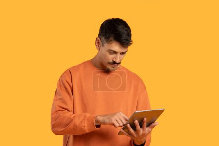 Photo for A man in an orange sweater is sitting while using a tablet. He appears focused on the screen, tapping and swiping. The background is blurred, emphasizing the man and his electronic device. - Royalty Free Image