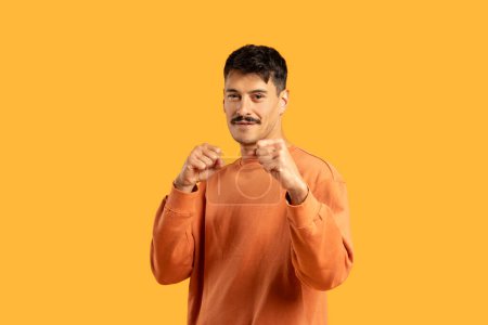 Ein selbstbewusster junger Mann reckt defensiv die Fäuste und trägt ein legeres orangefarbenes Sweatshirt. Er steht vor einem soliden gelben Hintergrund