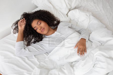 Foto de Una mujer está acostada en una cama con sábanas blancas limpias. Lleva un traje cómodo y parece estar relajada o descansando tranquilamente.. - Imagen libre de derechos
