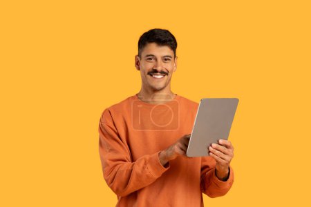 Foto de Un hombre con un suéter naranja es visto sosteniendo una tableta en sus manos. Él está mirando la pantalla atentamente, aparentemente comprometido con el contenido mostrado. El fondo es neutro, permitiendo el enfoque - Imagen libre de derechos