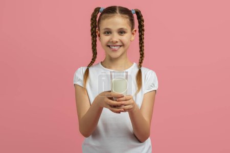 Teen girl holds a glass of milk, smiling pleasantly, isolated on a pink background, portraying a healthy lifestyle concept