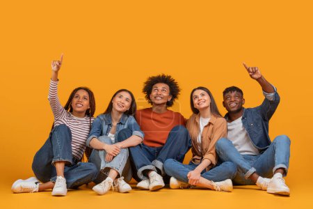 An international group of friends joyfully pointing upwards, showcasing unity and aspiration in a multiethnic, multiracial context, isolated on a yellow background