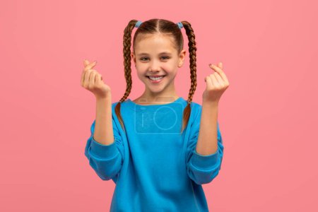 Téléchargez les photos : Une jeune fille joyeuse avec des cheveux tressés dans un haut bleu, souriant et faisant l'amour geste avec les doigts sur un fond rose - en image libre de droit