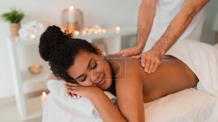Téléchargez les photos : Une image en gros plan d'une dame noire profitant d'un massage des épaules apaisant dans un spa, avec un accent sur les mains du thérapeute et une atmosphère relaxante - en image libre de droit