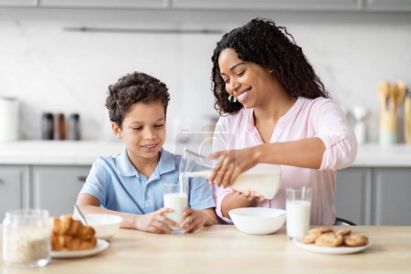 A caring African American mother pours milk for her son at the breakfast table, highlighting family support and kitchen interactions