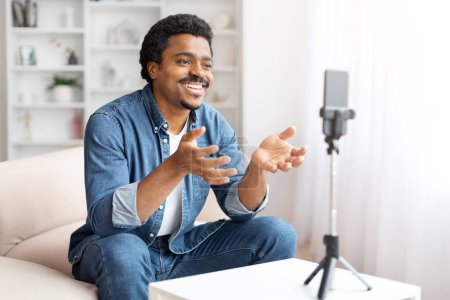 Lächelnder schwarzer Mann in Jeanshemd Blogger, der einen Videoblog auf einem Smartphone aufzeichnet, das auf einem Stativ in einem hellen Wohnzimmer montiert ist