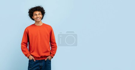 Foto de Relajado chico afroamericano con una sonrisa sutil, cómodamente vestido con suéter naranja y jeans, de pie con las manos en los bolsillos sobre un fondo azul aislado, espacio para copiar - Imagen libre de derechos