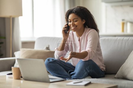 Foto de Cheerful young black woman engaging in a pleasant conversation over her smartphone at home, with an open laptop and a warm cup of coffee on the table - Imagen libre de derechos