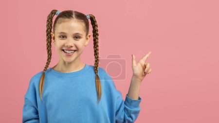 Téléchargez les photos : Joyeux jeune fille avec des cheveux tressés pointant vers le côté à l'espace de copie, portant un pull bleu sur un fond rose - en image libre de droit