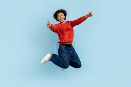 Foto de La imagen captura a un hombre afroamericano zoomer a mitad de salto, pulgares hacia arriba con una expresión extática, mostrando altos espíritus y libertad, contra un entorno aislado azul - Imagen libre de derechos
