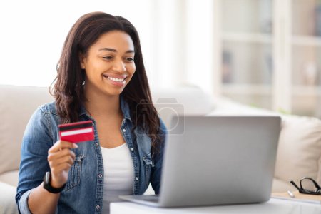 Photo for African American woman is shown holding a credit card in one hand and a laptop in the other. She appears to be engaged in online shopping or financial transactions - Royalty Free Image