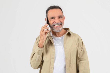Foto de Un hombre mayor está parado al aire libre mientras habla por un teléfono celular. Parece estar involucrado en una conversación mientras hace gestos con su mano libre.. - Imagen libre de derechos